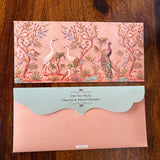 A peach-colored personalized money envelope featuring a detailed illustration of cranes, a peacock, and pink blossoms, displayed above its matching scalloped envelope.