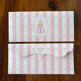 Two pink and white striped baby shower envelopes with bow patterns and a central illustration of a princess, displayed on a dark wood background.
